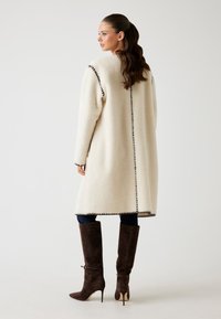 Cappotto in pelliccia sintetica color crema con cuciture scure a contrasto, lungo fino al ginocchio, dotato di maniche lunghe e taglio dritto, abbinato a stivali marroni sopra il ginocchio.