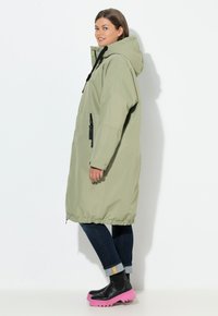 Lange lichtgroene waterdichte parka met een capuchon, zwarte trekkoorden en ritszakken. Draagt over donkere jeans en zwarte laarzen met roze zolen.