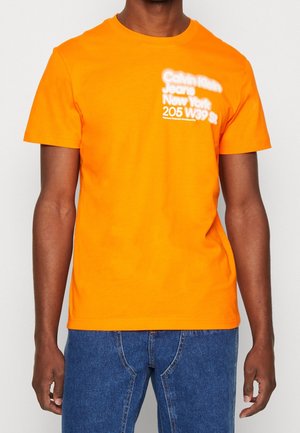 Mann trägt ein leuchtend oranges T-Shirt mit kurzen Ärmeln und weißem Text auf der Brust sowie blaue Jeans, stehend vor einem einfachen Hintergrund.
