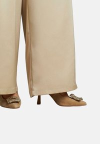 Pantalons larges beiges associés à des talons aiguilles beiges, avec un détail décoratif en boucle en strass sur le bout.