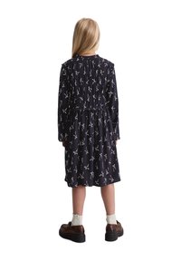 Marine jurk met een geraffineerde lijfje met witte strikpatronen, lange mouwen en een uitlopende rok, gecombineerd met bruine schoenen en crèmekleurige sokken.