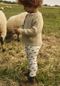 Beiger Rippenpullover, weiße Leggings mit blauen und braunen Tiermotiven, braune Knöchelstiefel. Kind steht auf einem Feld mit Schafen im Hintergrund.