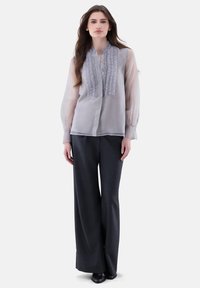Helles graues, durchsichtiges Blusenshirt mit Rüschen und Knopfschluss, kombiniert mit dunklen, weit geschnittenen Hosen und schwarzen Schuhen, die ein kontrastierendes Outfit bilden.