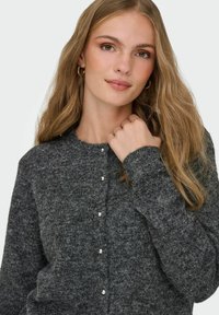 Ung kvinna med långt blont hår, iförd guldbaserade hoop-örhängen och en mörkgrå knappad stickad cardigan, som håller i kragen med ena handen.