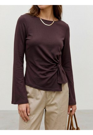 Mujer que lleva una camiseta de manga larga oscura con detalle de nudo lateral, pantalones beige, collar dorado y sostiene un bolso marrón.