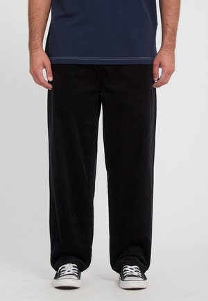 Homme portant un pantalon en velours côtelé noir ample, une chemise bleu marine et des baskets lacées noires et blanches, debout devant un fond uni.