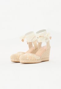 Kile espadriller med en vevd stråbase, kremfarget stoffoverdel og pastellfarget blomsterbroderi. Har snøring rundt ankelen og en rund tå.