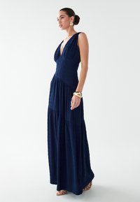 Marineblaues Maxikleid mit tiefem V-Ausschnitt aus gekrepptem Stoff. Eng anliegender Miederteil mit Empire-Taille und gestufter Rock. Akzent durch goldenes Armreif-Armband.
