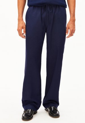 Homme portant un pantalon à cordon de serrage bleu marine et des mocassins à pampilles noirs debout devant un fond blanc.