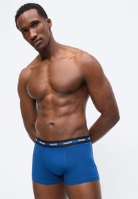 Μυώδης άνδρας που στέκεται με τα χέρια πίσω από την πλάτη, φοράει μπλε εσώρουχα Hugo boxer, με απλό ανοιχτό φόντο.