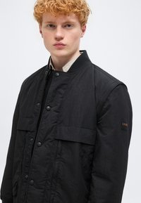 Νεαρός άνδρας με σγουρά κόκκινα μαλλιά, φορώντας μαύρο σακάκι με κουμπιά και τσέπες με καπάκι, στέκεται μπροστά σε απλό ανοιχτό φόντο.