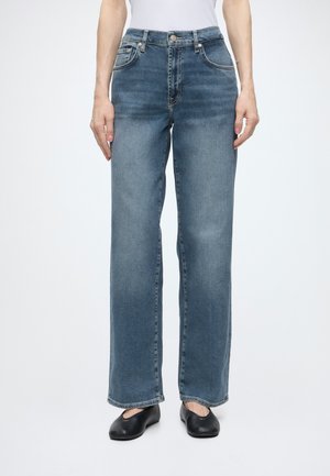 Frau trägt blaue gerade geschnittene Jeans, weißes Top und schwarze flache Schuhe, steht mit entspannt an den Seiten hängenden Armen.
