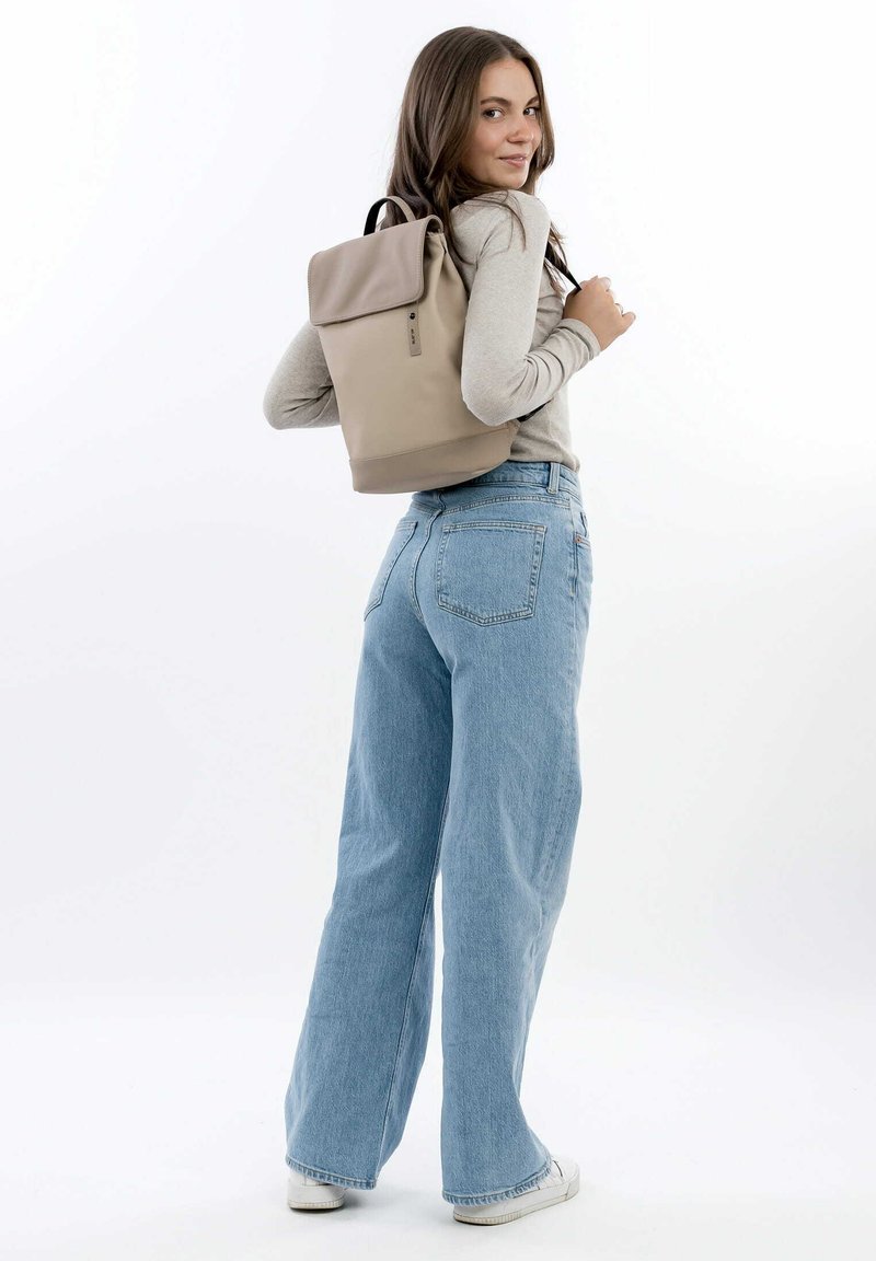 Zaino beige a roll-top con texture liscia, hardware nero e spallacci regolabili; indossato con jeans larghi a vita alta blu chiaro.