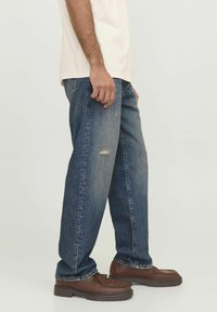Blue denim jeans with a loose fit, featuring a distressed knee detail, paired with brown leather shoes. The denim has a faded texture.