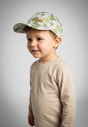 Junger Junge lächelt, trägt ein hellbeiges Langarmshirt und eine weiße Mütze mit grünen Dinosauriermustern.
