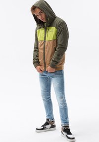 Huvan jacket med grön överdel, brun underdel och frontdragkedja. Har elastiska ärmslut och två sidofickor. Bärs med slitna jeans och sneakers.