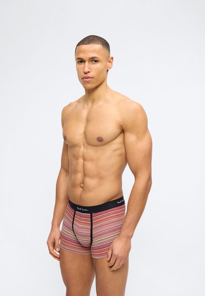 Μυώδης άνδρας όρθιος, φορώντας πολύχρωμα ριγωτά μποξεράκια με μαύρη μέση, σε απλό λευκό φόντο.