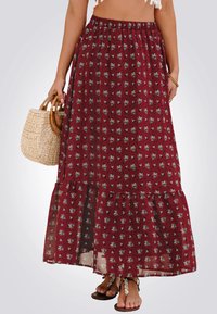 Jupe longue bordeaux avec de petits motifs floraux, ourlet à volants, taille élastique, fabriquée en tissu léger ; portée avec des sandales marron et un panier tressé.