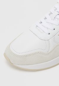 Bílé tenisky s hladkým koženým svrškem, šedými semišovými akcenty, perforovanými detaily a plochými bílými tkaničkami. Zaoblený design špičky.