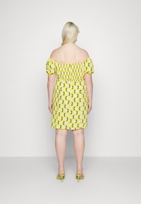 Vestido off-the-shoulder amarelo com padrão floral, corpete franzido, mangas curtas bufantes e saia ajustada. Combinado com mules de salto alto verdes.