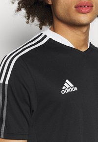Osoba v černém sportovním tričku Adidas s bílými třemi pruhy na ramenou a logem Adidas na hrudi.