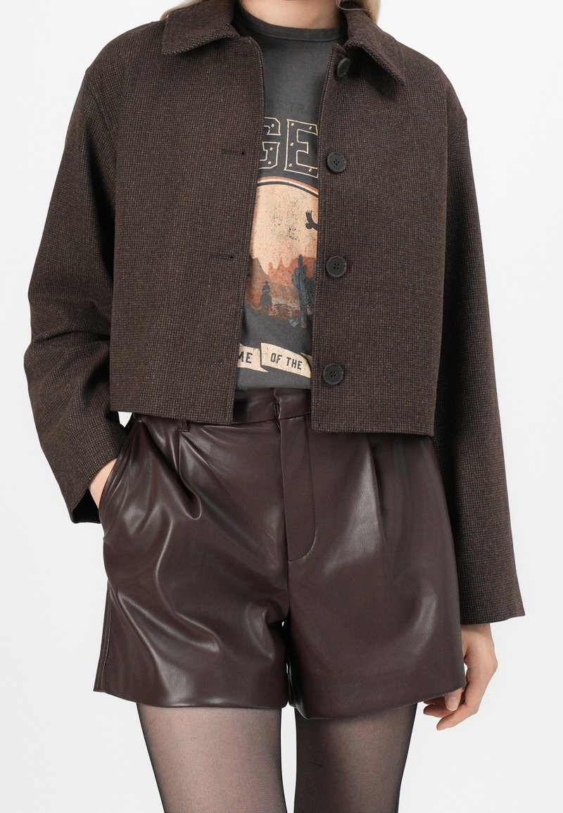 Veste marron courte à boutons portée sur un t-shirt noir graphique, associée à un short en cuir marron foncé taille haute et des collants noirs transparents.