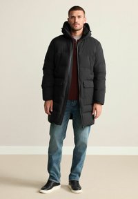 Schwarze Steppjacke mit Kapuze, Reißverschluss und zwei Fronttaschen; getragen über einem burgunderfarbenen Hemd und blauen Jeans; strukturierter Stoff.