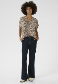 Blus med kort ärm i leopardmönster i beige och brun, kombinerad med mörkblå utsvängda jeans. Modellen har på sig ljusa slip-on-skor.