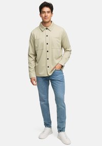 Lys beige skjorte med knapper og krage, med to brystlommer, iført over blå jeans og hvite sneakers. Glatt stoff, avslappet passform.
