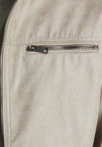 Bežová semišová bunda s kapsou na zip se stříbrným kováním. Měkká textura a čisté detaily šití jsou viditelné.