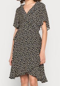 Robe noire avec un décolleté en V et des manches volantées, ornée d'un motif en cœur doré et blanc sur un tissu léger. Ourlet évasé.