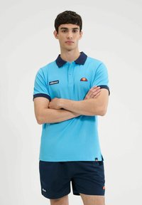 Polo di colore azzurro chiaro con colletto e dettagli delle maniche in navy; presenta un piccolo logo arancione e giallo e un'etichetta nera con "ellesse."