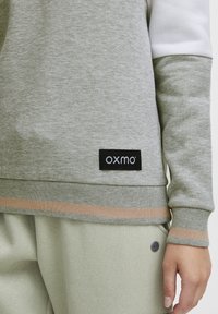 Sivi dukserica s bijelim rukavima, s crnim „OXMO“ logom i horizontalnom bež prugom na rubu. Mekana, teksturirana tkanina.