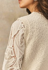 Kremowy sweterek na plecach z teksturowanym wzorem, w zestawie z bluzką z koronkowymi detalami na rękawach.