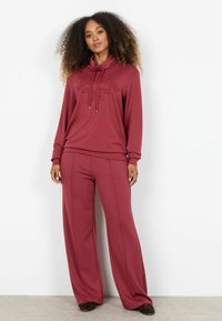 Hnedá mikina s kapucňou a reliéfnym textom, kombinovaná s nohavicami s širokými nohavicami v rovnakej farbe. Mäkká tkanina, uvoľnený strih, minimálne detaily.