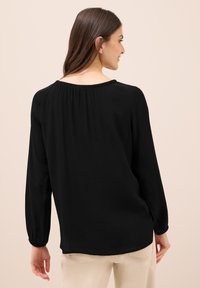 Blusa nera a maniche lunghe con scollo rotondo e dettaglio dietro arricciato. Il tessuto è liscio, con una vestibilità comoda e polsini elastici.