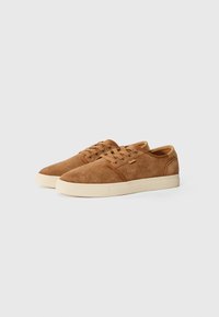 Zapatillas de ante marrón con una textura suave, suela de goma beige, puntera redonda y acentos laterales perforados; diseño clásico con cordones.
