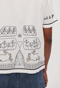 Camisa blanca de manga corta con diseños bordados en negro y marrón de conejos, plantas y una valla, con un trabajado diseño en el dobladillo.
