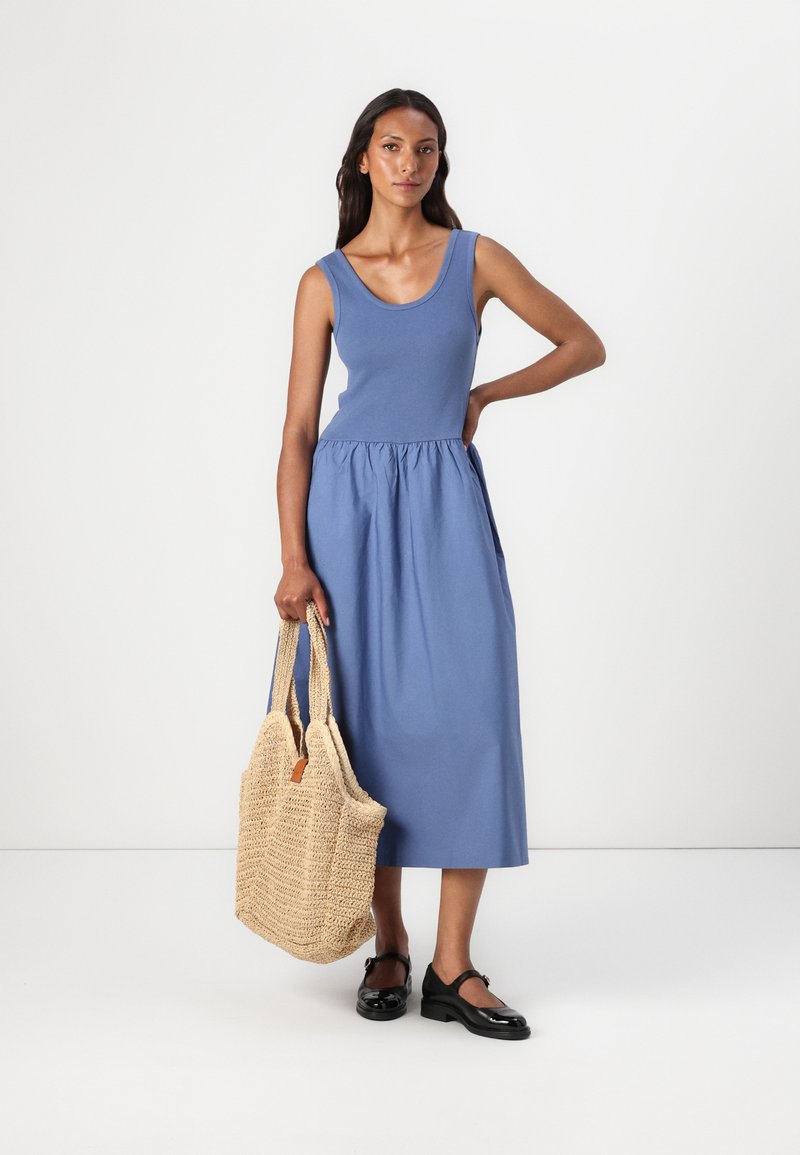 Ärmelloses blaues Baumwollkleid mit geraffter Taille, kombiniert mit einer gewebten beigefarbenen Tragetasche und schwarzen Schuhen. Einfaches, klares Design.
