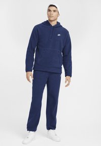 Felpa in pile blu navy con tasca frontale e cappuccio con lacci, abbinata a pantaloni tuta tapered coordinati e sneakers bianche.