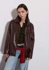 Giovane donna con capelli castani lisci, che indossa una giacca di pelle marrone scuro, una maglietta nera, jeans azzurri chiari e un maglione a righe rosso annodato in vita.