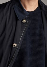 Navy blue jacket with a buttoned collar and silver snap buttons, layered over a textured navy sweater with a ribbed neckline.