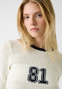 Licht crèmekleurige trui met opengewerkt breisel, zwarte kraag en een opvallende gestikte "81" aan de voorkant. Zachte textuur, losse pasvorm.