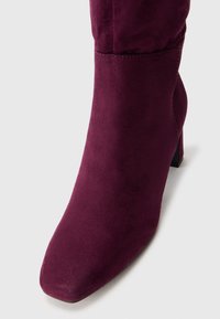 Botine din suede burgund cu vârf pătrat și toc conic, având o textură netedă și detalii de cusătură minime.