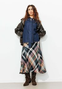 Blouse en denim avec un nœud à l'avant, associée à une jupe maxi à carreaux multicolores. Veste bomber en cuir marron et bottes marron jusqu'au genou.