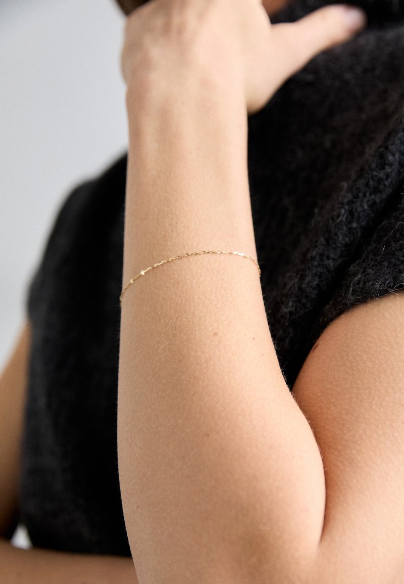 Zlatna lančić narukvica na podlaktici, s delikatnim karikama i glatkom teksturom, nošena uz tamnu, teksturiranu tkaninu.