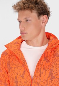 Giacca arancione con un motivo a linee geometriche, colletto alto e dettagli con zip. Indossata sopra una camicia di colore chiaro, mette in mostra il suo design vivace.