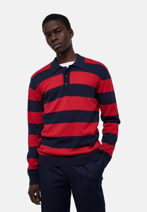Jeune homme portant un polo à manches longues avec de larges rayures horizontales rouges et bleu marine, associé à un pantalon foncé, debout avec une main dans la poche.