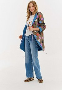 Zielono-niebieska kurtka w kropki z różowymi sznurkami, biała koszulka z grafiką, szerokie niebieskie dżinsy i buty w panterkę.