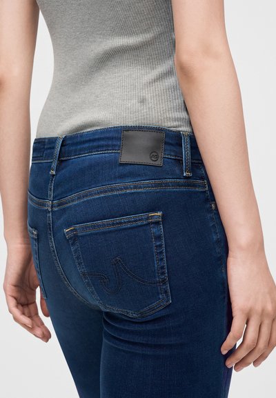 Κοντινό πλάνο ενός ατόμου που φορά σκούρα μπλε τζιν και γκρι ριμπ αμάνικη μπλούζα, με οπίσθιες τσέπες και μαύρο δερμάτινο λογότυπο.